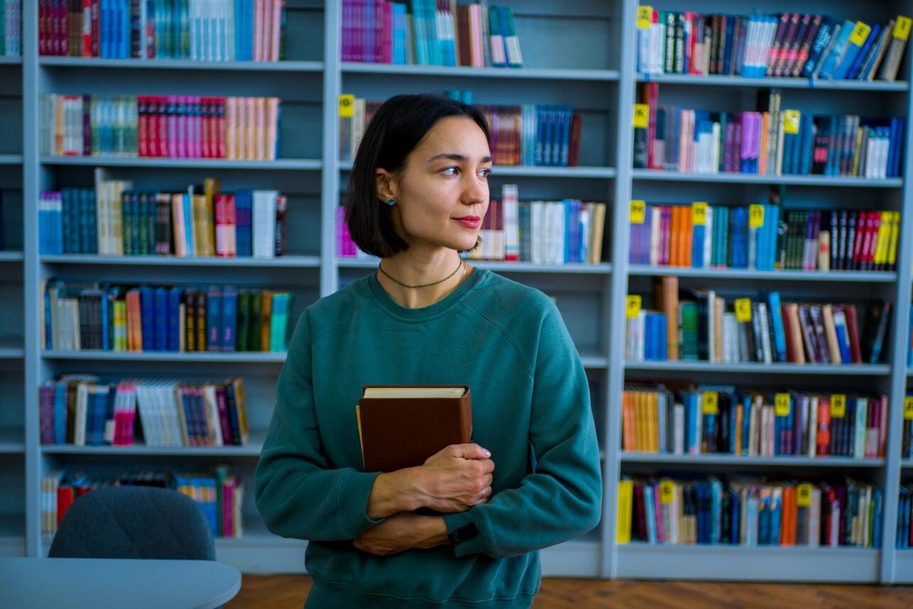 Молодая женщина в зеленом свитшоте с книгой на фоне книжных полок, смотрит в сторону.